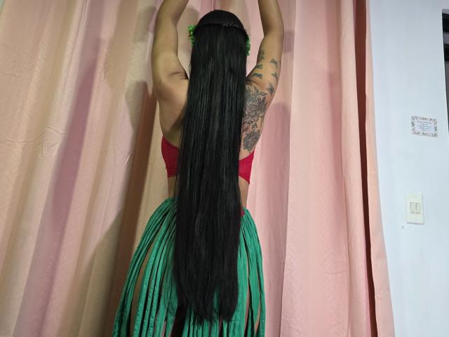 Yurimy with long black hair, red bra, and green skirt, arms raised, tattooed right arm, standing against pink curtains, back view, showing her ass.