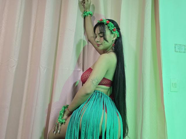Yurimy with long black hair, wearing a red lace bra, green floral headband, and teal grass skirt, touching a pink curtain, showing her tattooed arm and curvy ass.
