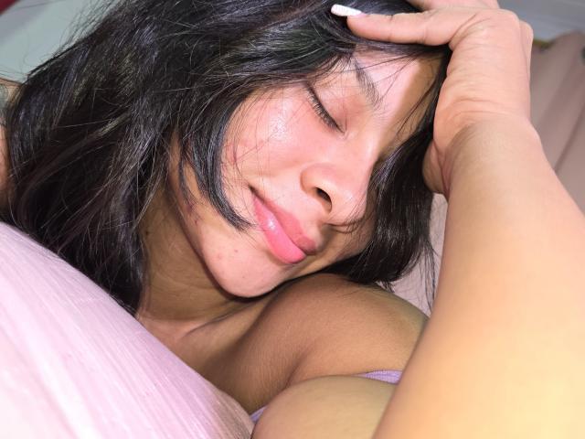 Yurimy with closed eyes, black hair, and pink lips, resting her head on a pink pillow, hand in hair, showing her tits slightly. Her skin is tan and shiny.