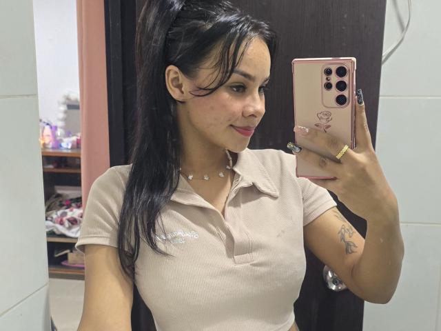 Yurimy with black hair in a ponytail, wearing a beige polo, holding a pink phone, smiling, showing a tattoo on her arm, in a bathroom mirror selfie.