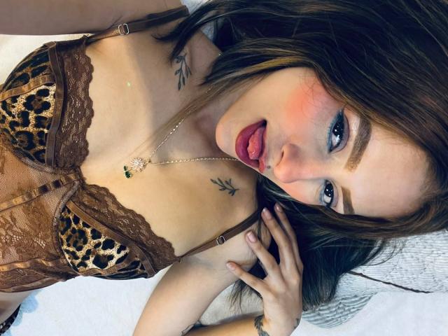AfroditaWild, com lingerie marrom estampada de leopardo, língua para fora e olhando para cima. Ela tem olhos azuis, tatuagens no pescoço e clavícula, e um colar de ouro com um pingente verde..