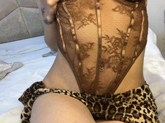 AfroditaWild vestindo um macacão de renda marrom com estampas florais, mostrando sua virilha e uma saia de estampa de leopardo puxada para baixo, expondo sua vulva. Seu braço tatuado está visível..