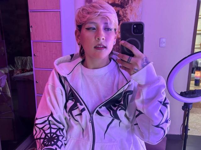DannyRouds met kort roze haar, een witte hoodie met zwarte spinnenwebpatronen, die een telefoon vasthoudt, paars en roze verlichting, een ring aan de vinger, spiegelreflectie, een ringlamp aan de rechterkant..