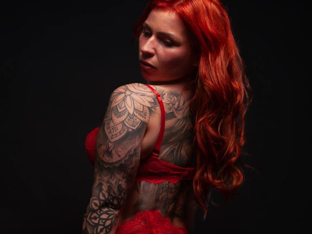 Chrissi69, aantrekkelijk met rood haar, draagt een rode kantbh, kijkt om en toont haar gedetailleerde getatoeëerde arm, en laat haar sexy, ronde billen zien. De donkere achtergrond benadrukt haar vurige, verleidelijke uitstraling..