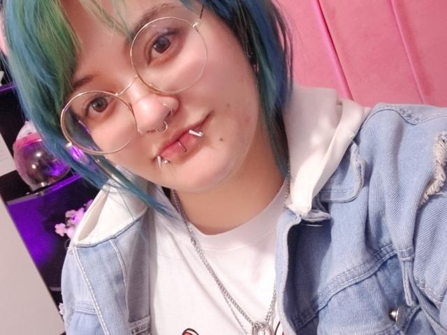 LuckyLeahh with blue-green hair, glasses, nose ring, lip piercings, and denim jacket over white shirt, showing a cute, slightly mischievous smile.