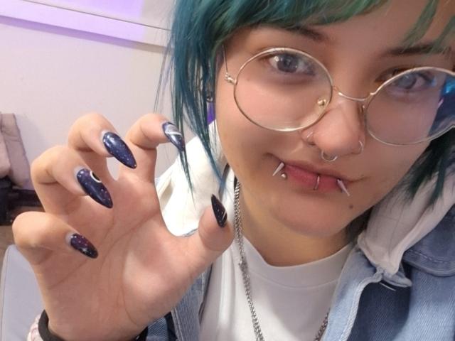 LuckyLeahh with teal hair, glasses, nose ring, lip piercings, and dark blue glitter nails, showing off her nails and smiling. She's wearing a white shirt and denim jacket.