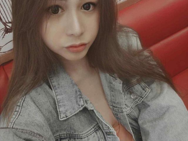 LovelyAya, con lunghi capelli castani, indossa una giacca di jeans, mostrando leggermente il décolleté, seduta in una cabina di pelle rossa, che guarda direttamente la fotocamera con un'espressione neutra..