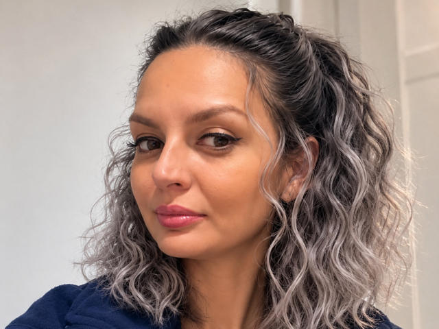 DouceAmelia heeft golvend, zilverkleurig haar in een rommelige staart, een bruine huid, subtiele make-up, roze lippen en donkere wenkbrauwen. Ze draagt een marineblauwe top en kijkt iets naar rechts..