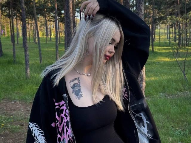 SerinaSindlinger has blonde hair, red lipstick, black tank top, black jacket with pink and white designs, and a tattoo on her right chest. She's standing in a forest.