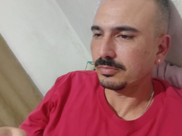MellyHomlly, con la cabeza calva, un bigote oscuro y pequeños aretes, vestida con una camisa roja y un collar delgado, mirando hacia un lado con una expresión neutra..