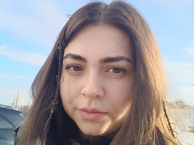 SamantaMais has brown eyes, a cute nose, and pouty lips, with long brown hair in braids, wearing a black jacket, against a blue sky.