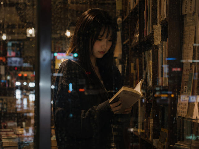 EmaVanhowe with dark, wavy hair, wearing a plaid sweater, reading a book in a dimly lit, book-filled store at night. Reflections on the window. Soft, moody lighting.