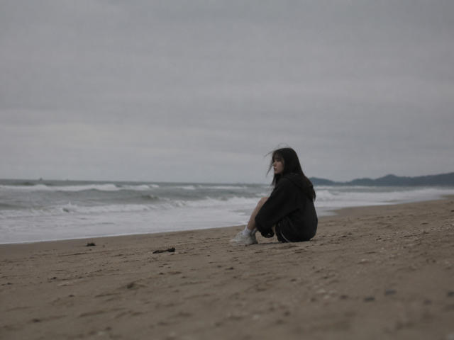 EmaVanhowe sits alone on a sandy beach, wearing a black hoodie and gray shoes, with her back to the camera, looking at the ocean.