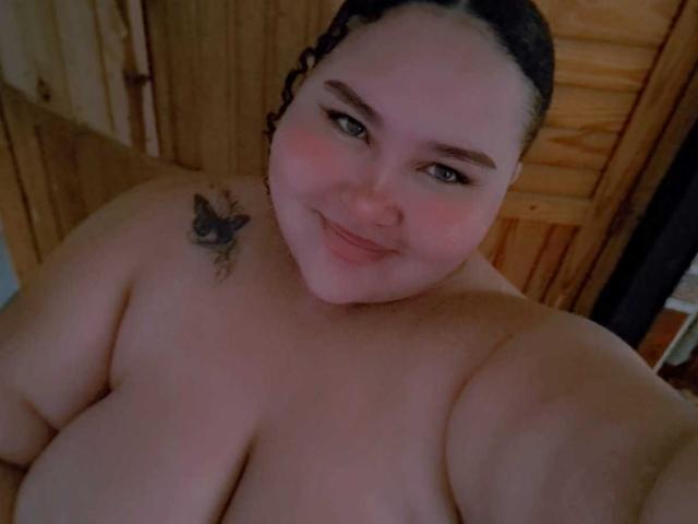 HurrenMatt, a curvy, topless woman with dark curly hair, a butterfly tattoo on her left shoulder, and a soft smile, showing her large, bare breasts.
