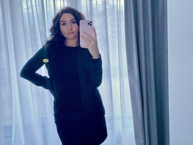 Dionisia with curly brown hair, wearing a black dress and jacket, holding a pink iPhone, taking a mirror selfie, showing her ass, standing with one hand on her hip.