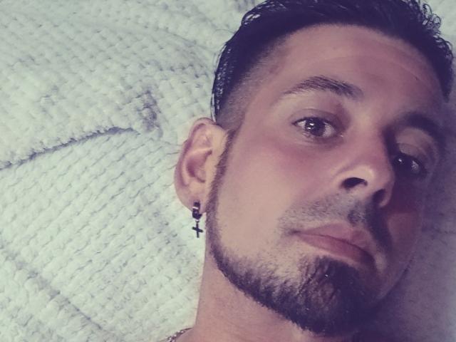 NinaetRicco hat einen kurzen, dunklen Bart, ein kleines Kreuz-Ohrring und ein leichtes Lächeln. Sie liegt auf einer weißen, strukturierten Decke. Ihre Augen sind braun und sie schaut in die Kamera..