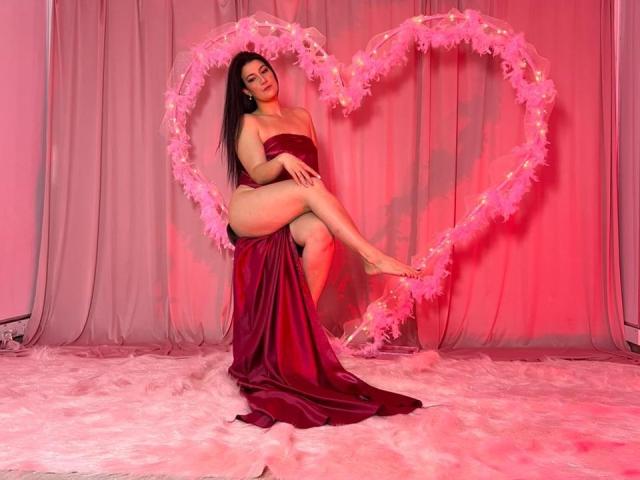 YessiSoto with long dark hair, wearing a red satin dress, sitting with one leg raised, in front of a pink heart made of feathers, on pink furry carpet.