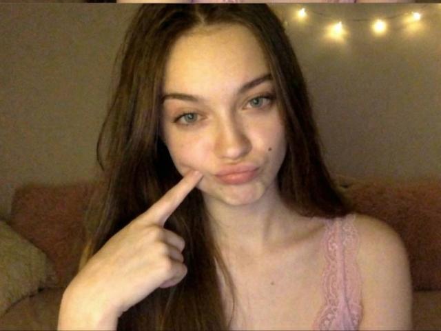 Diantrisy with long brown hair, pouty lips, and a cute face, pointing at her slightly smushed nose. Wearing pink lace, she's looking at the camera with a playful expression.