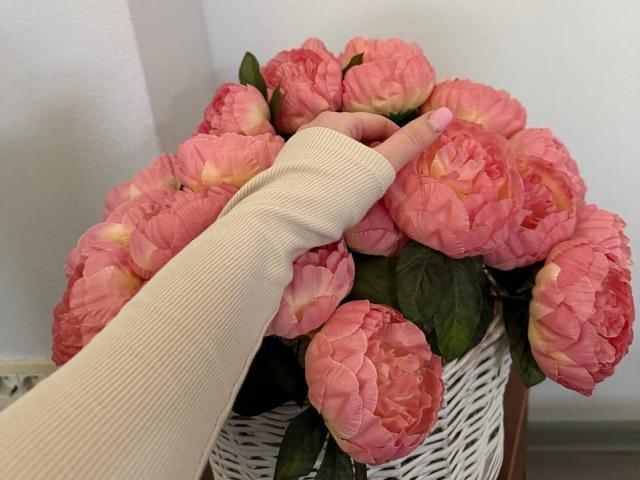La mano de Diantrisy, con un dedo pintado de rosa, toca suavemente un ramo de flores de peonía rosadas en una cesta de mimbre blanca..