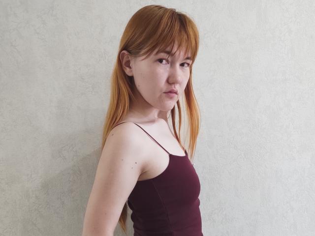 SheritaChieng has red hair, light skin, small breasts, and wears a maroon tank top, standing against a textured white wall, looking slightly to the side with a neutral expression.