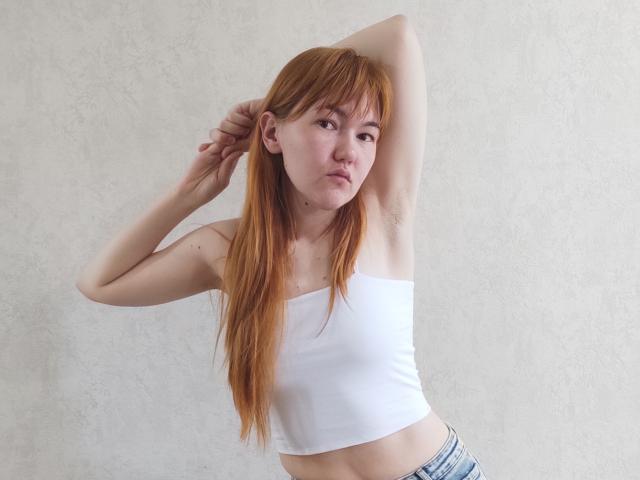 SheritaChieng heeft rood haar, een wit topje zonder bandjes, en blauwe jeans, met één arm omhoog, waarbij haar oksel zichtbaar is, en een frons op haar gezicht..