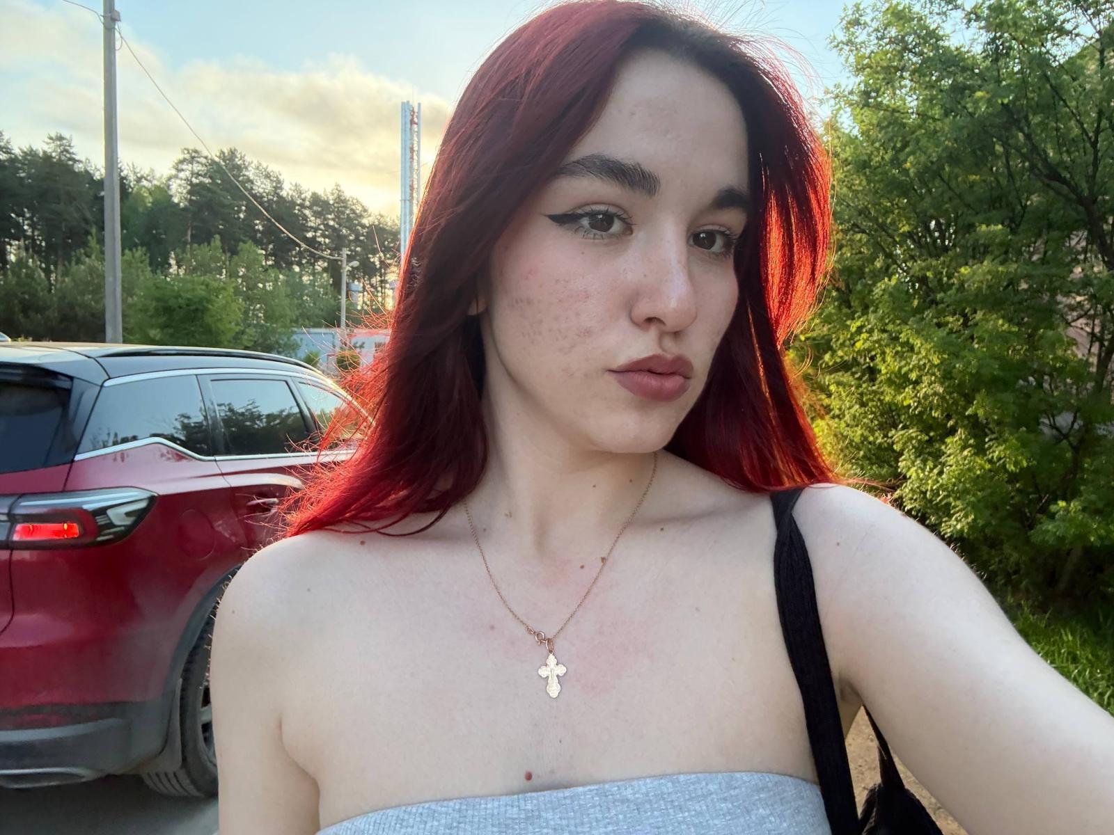 TheaWeemes has red hair, pale skin, and small tits. She's wearing a strapless gray top, black shoulder strap, and a gold necklace with a small cross. Outside, red car.