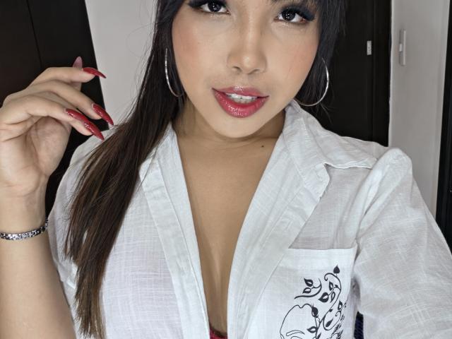 Heyly with shiny red lips, long dark hair, and red nails, wearing a white shirt showing her tits, hoop earrings, and a bracelet, looking at the camera.