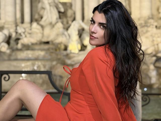 ZoeeMar with long black hair, wearing an orange dress, sitting with her back to the camera, looking over her shoulder, showcasing her bare ass and smooth skin.