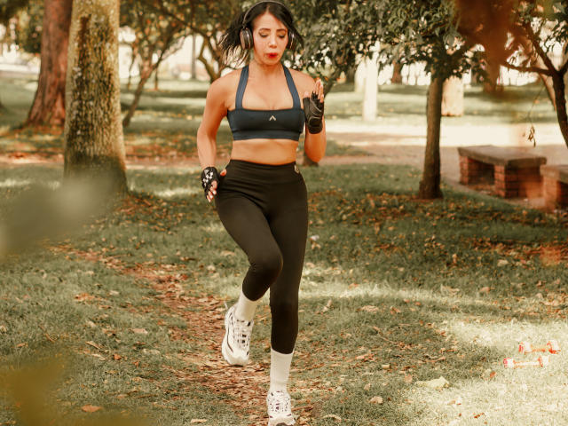 HelenHard, running in a park, wearing a black sports bra, black high-waisted leggings, white socks, and white sneakers, with black fingerless gloves, and her tits are visible.