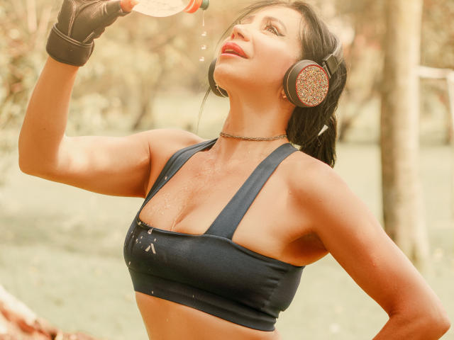 HelenHard drinking water, wearing a black sports bra, showing her tits, with sweat on her chest, black gloves, and large headphones, in a sunny outdoor setting.