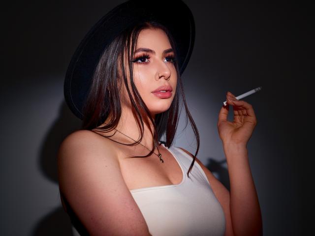 VanessaClose with long brown hair, wearing a black hat and white tank top, holding a cigarette, has pink lipstick and dark eyeliner, with a small cross necklace.