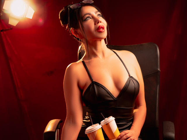 HelenHard with dark hair in a bun, wearing a black leather dress, holding drinks, sitting in a chair, looking up with red lips, sunglasses on head, red background.