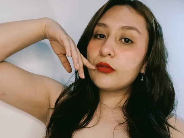 NathashaBell con lápiz labial rojo, cabello ondulado oscuro y una ligera expresión de labios fruncidos, tocándose el labio con el dedo, usando un collar y aretes, sobre un fondo blanco..