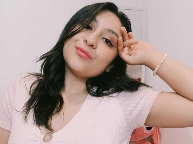 NathashaBell, con i capelli neri, una camicia rosa e rossetto rosa, che si tocca la fronte, sorridendo leggermente, indossa un braccialetto di perle e mostra il suo sedere carino e formoso..