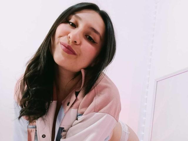 NathashaBell, com cabelos longos e pretos, jaqueta rosa e branca, e calcinha de renda branca, sorrindo com batom rosa, seus seios parcialmente visíveis, inclinada para frente, parecendo fofa e sexy..