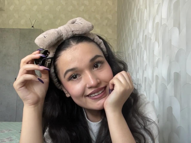 Meaghan Grigalonis tem cabelos longos e escuros, usa uma tiara rosa fofa com orelhas, está sorrindo, segurando um telefone no ouvido, tem unhas roxas, veste uma camisa branca e tem um piercing no nariz..