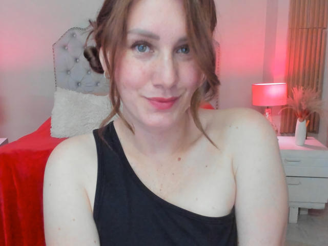 KatyaRogersX with fair skin, brown hair in messy buns, blue eyes, black one-shoulder top, slight smile, red lips, small moles on chest, in a bedroom with red and pink lights.