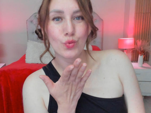 KatyaRogersX, puffy pink lips, blue eyes, brown hair in messy bun, black strapless top, hand up, pale skin, bedroom with red bed, white nightstand, red lamp.