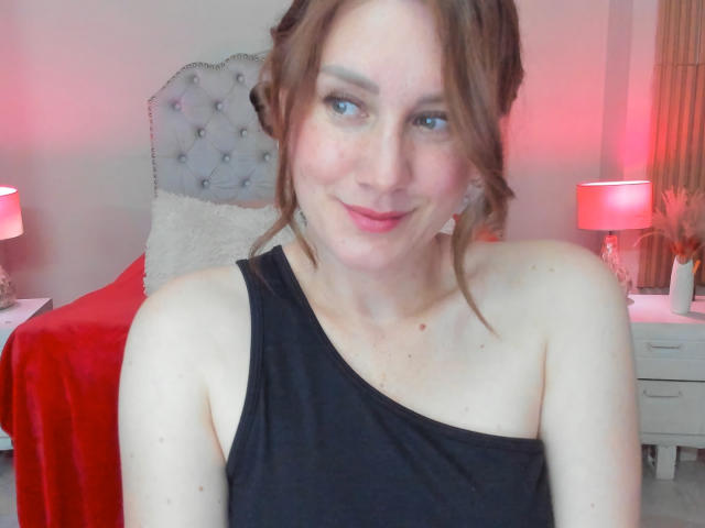 KatyaRogersX, with fair skin, blue eyes, and brown hair in a messy updo, wearing a black one-shoulder top, smiling softly at the camera. Her tits are small.
