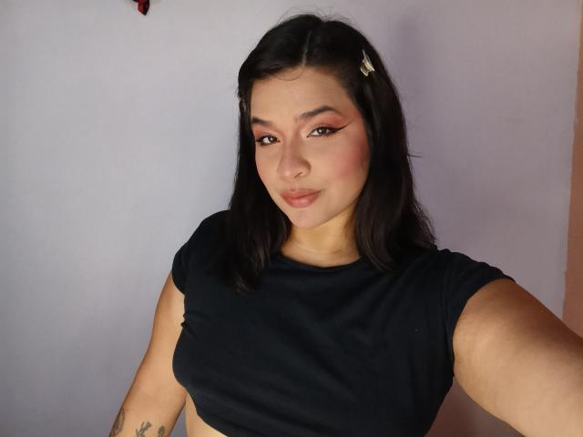 AntonelaPark with black hair, pink eyeshadow, and a small hair clip, wearing a black top, showing her tits and a tattoo on her right arm, smiling at the camera.