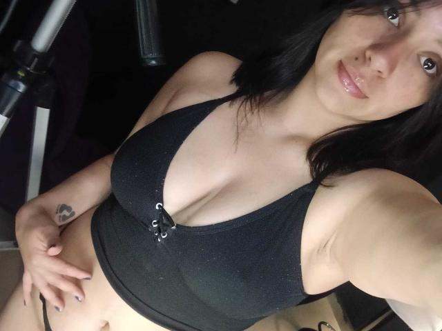 ZaharaViscon com lábios brilhantes, blusa de alça preta que mostra seios grandes, cabelo preto, mão na cintura, tatuagem no pulso esquerdo, em pé em um quarto escuro, olhando para a câmera..