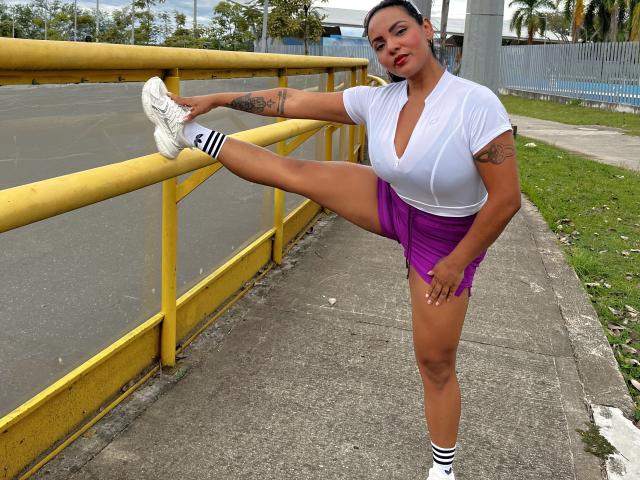 JessicaFiore, com seios grandes, shorts roxos, camiseta branca e tênis brancos, esticando uma perna contra um corrimão amarelo, mostrando tatuagens e usando meias listradas..