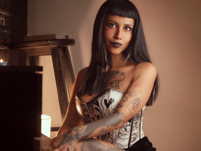 NyxVega with long black hair, dark lipstick, tattoos on arms and chest, white and black corset, sitting in dim light, candle on left, wood ladder in background.