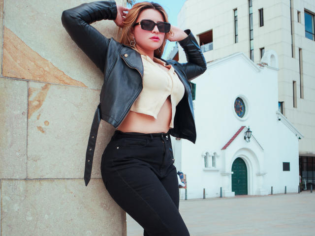 SaraWestt with sunglasses, black leather jacket, beige crop top, and black jeans, leaning against a stone wall, hands in hair, showing her tits and ass. Urban street setting.