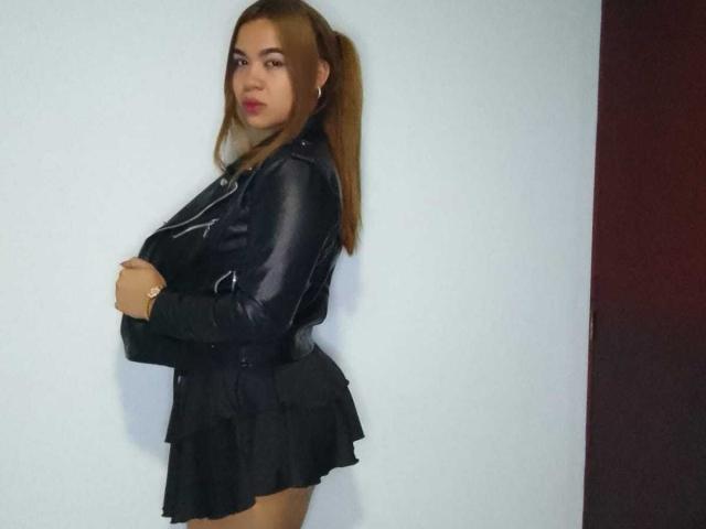 KieraRoss stands with arms crossed, wearing a black leather jacket and ruffled black skirt, her brown ponytail over her shoulder, small tits, light skin, and red lipstick.