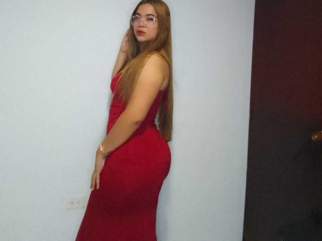 KieraRoss with long blonde hair, glasses, and a tight red dress, showing her ass and a gold bracelet, standing against a light blue wall.