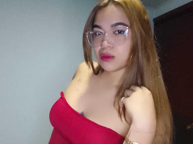 KieraRoss with long brown hair, red strapless top, clear glasses, and pink lipstick, showing her tits and a small tattoo on her right shoulder.