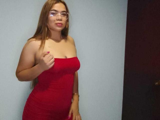 KieraRoss with long brown hair, glasses, and red strapless dress, showing her tits with right hand, smiling, against a light blue wall, right side dark door.