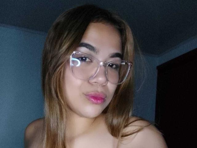 KieraRoss with glossy pink lips, clear glasses, and brown hair, slightly winking, showing her bare shoulders, has a cute and confident expression.