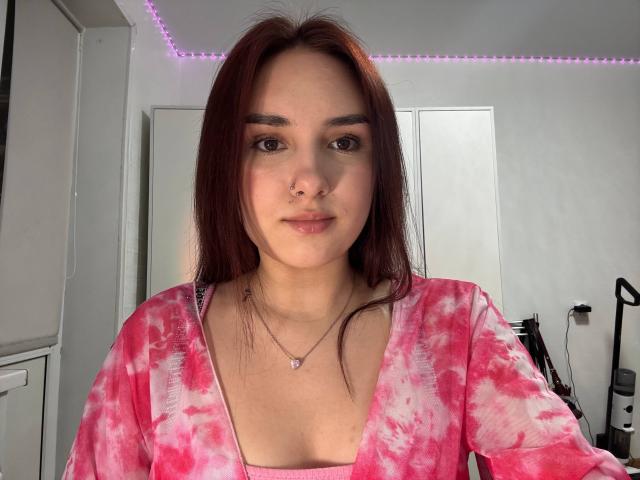 LashondaAllhands with red hair, nose ring, pink tie-dye shirt, small tits, and pink necklace, looking at camera in bright room.